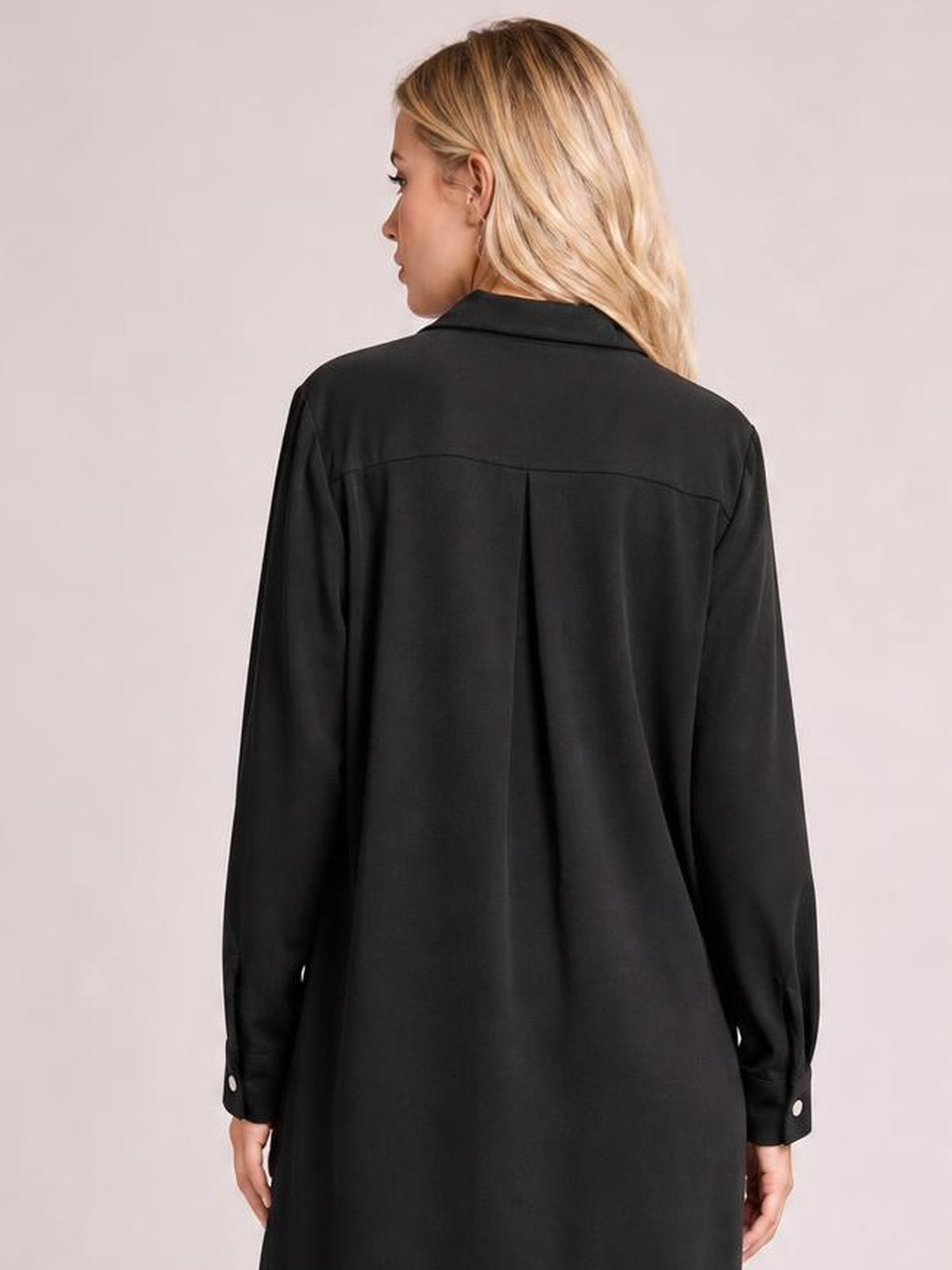 A model poses from the back in a black shirt dress with a pointed collar, long sleeves, buttoned cuffs, back yoke seam, and center pleat.