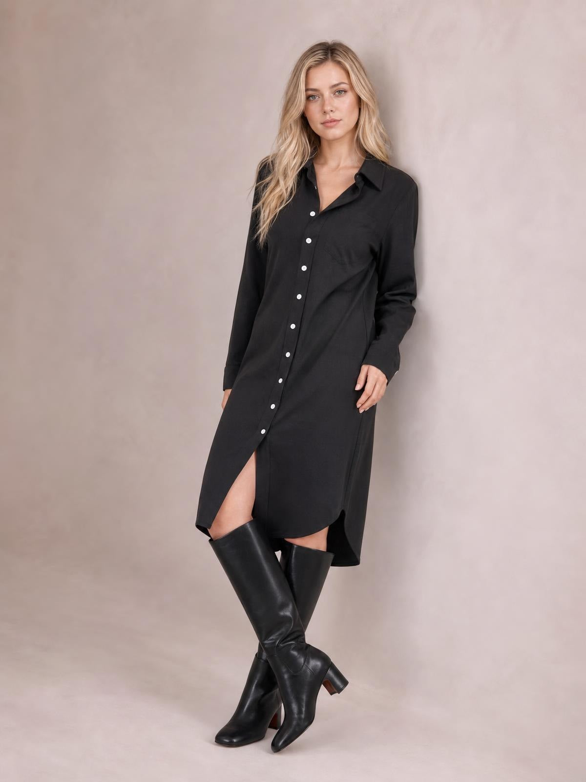 A model poses with crossed legs in a black shirt dress featuring a collared button-down front, long sleeves, relaxed silhouette, and curved midi-length hem.