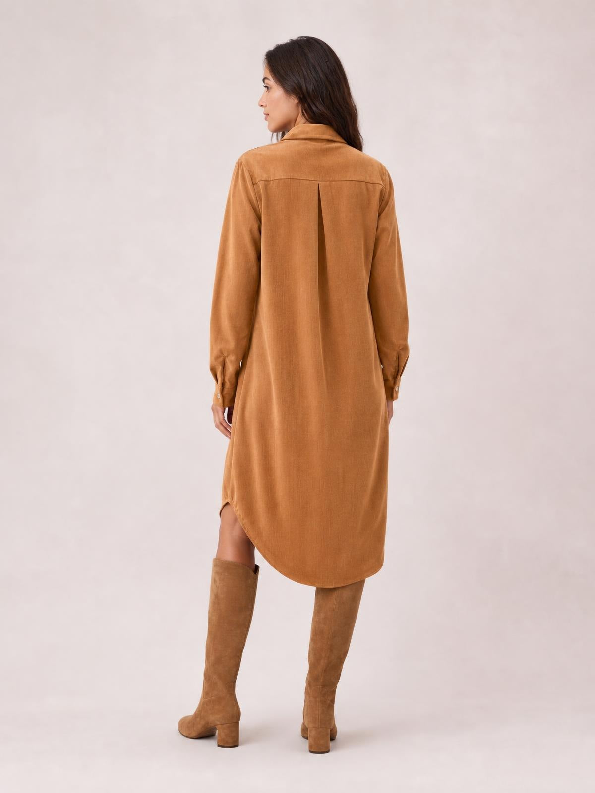 Model poses with her back turned, showing a camel corduroy shirt dress with a collared neckline, long sleeves, center-back pleat, and curved midi hem.