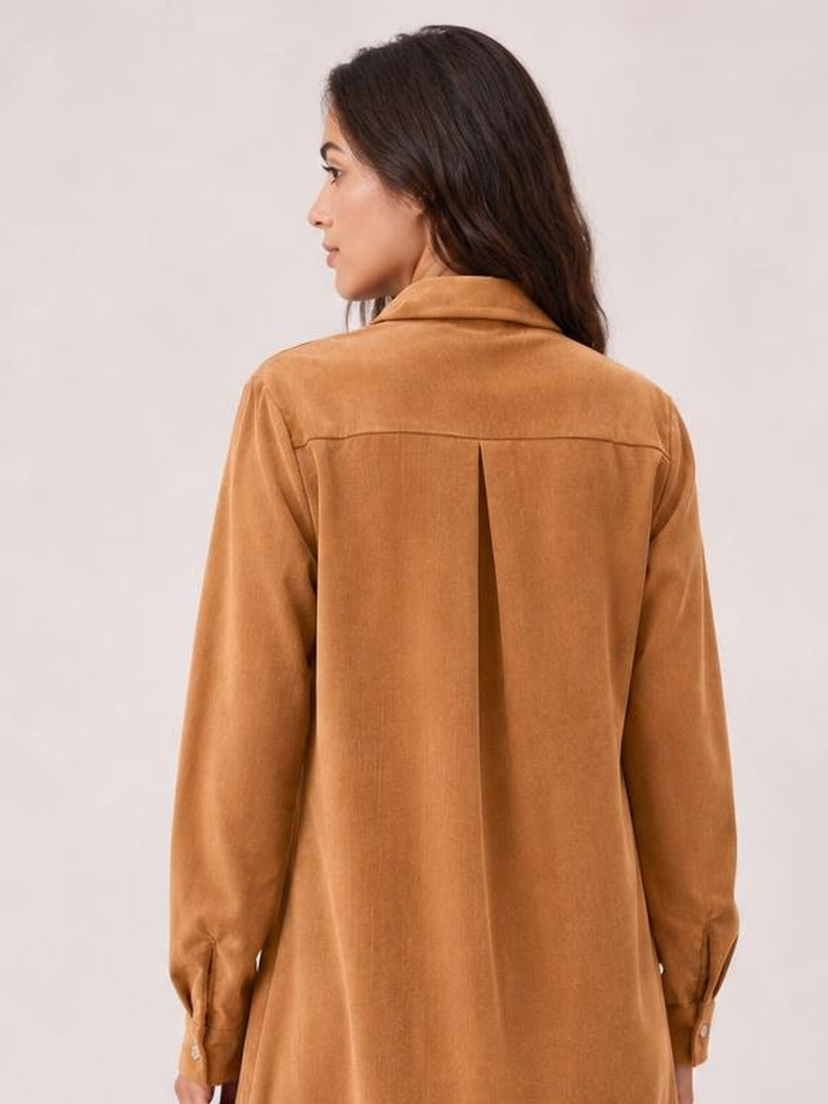 A model shows the back of a camel corduroy shirt dress with a collar, long sleeves, back yoke, center pleat, and relaxed straight silhouette.