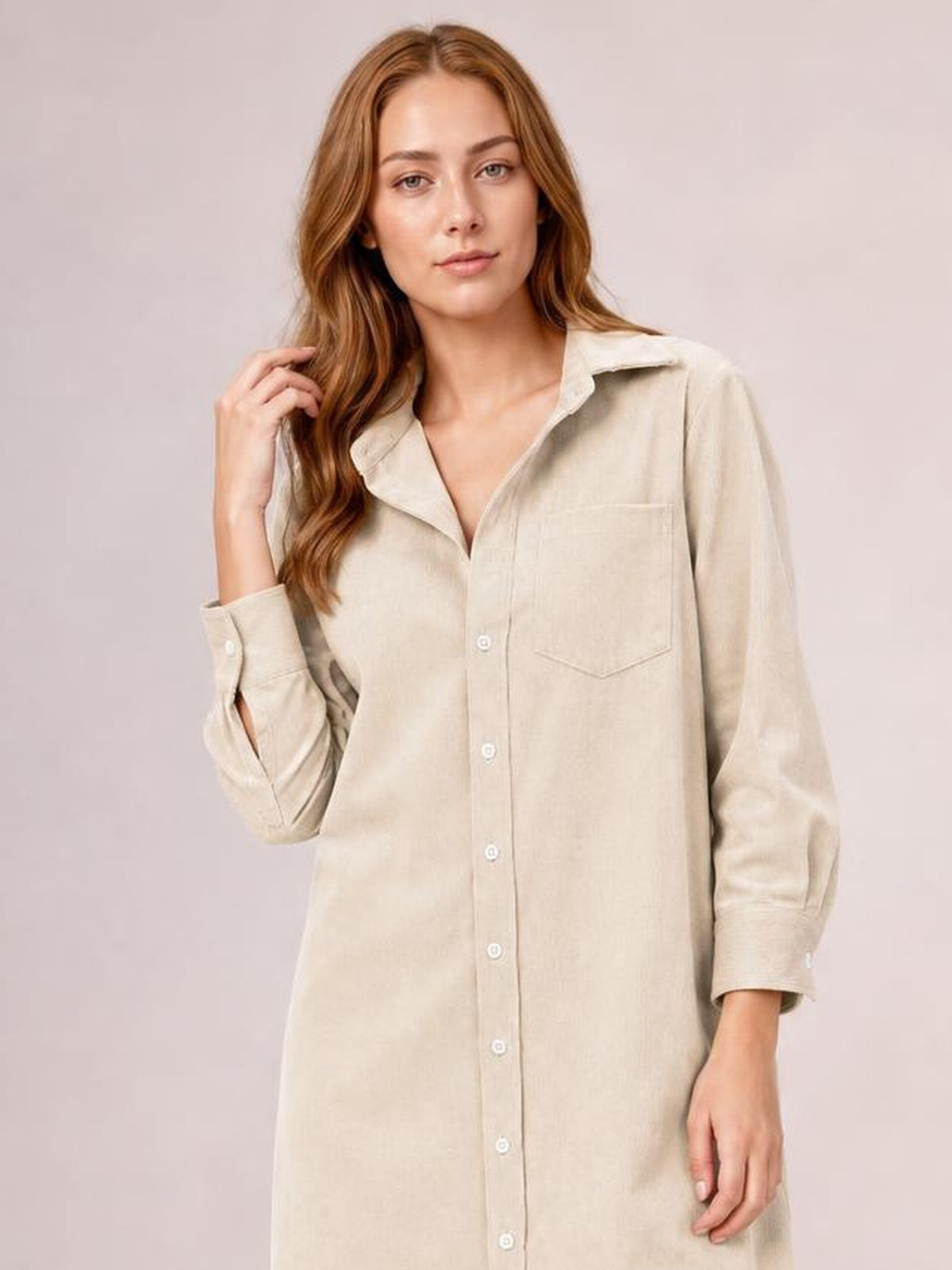 Model poses with one hand in her hair wearing a beige corduroy shirt dress with a button-down front, pointed collar, long sleeves, chest pocket, and relaxed straight silhouette.