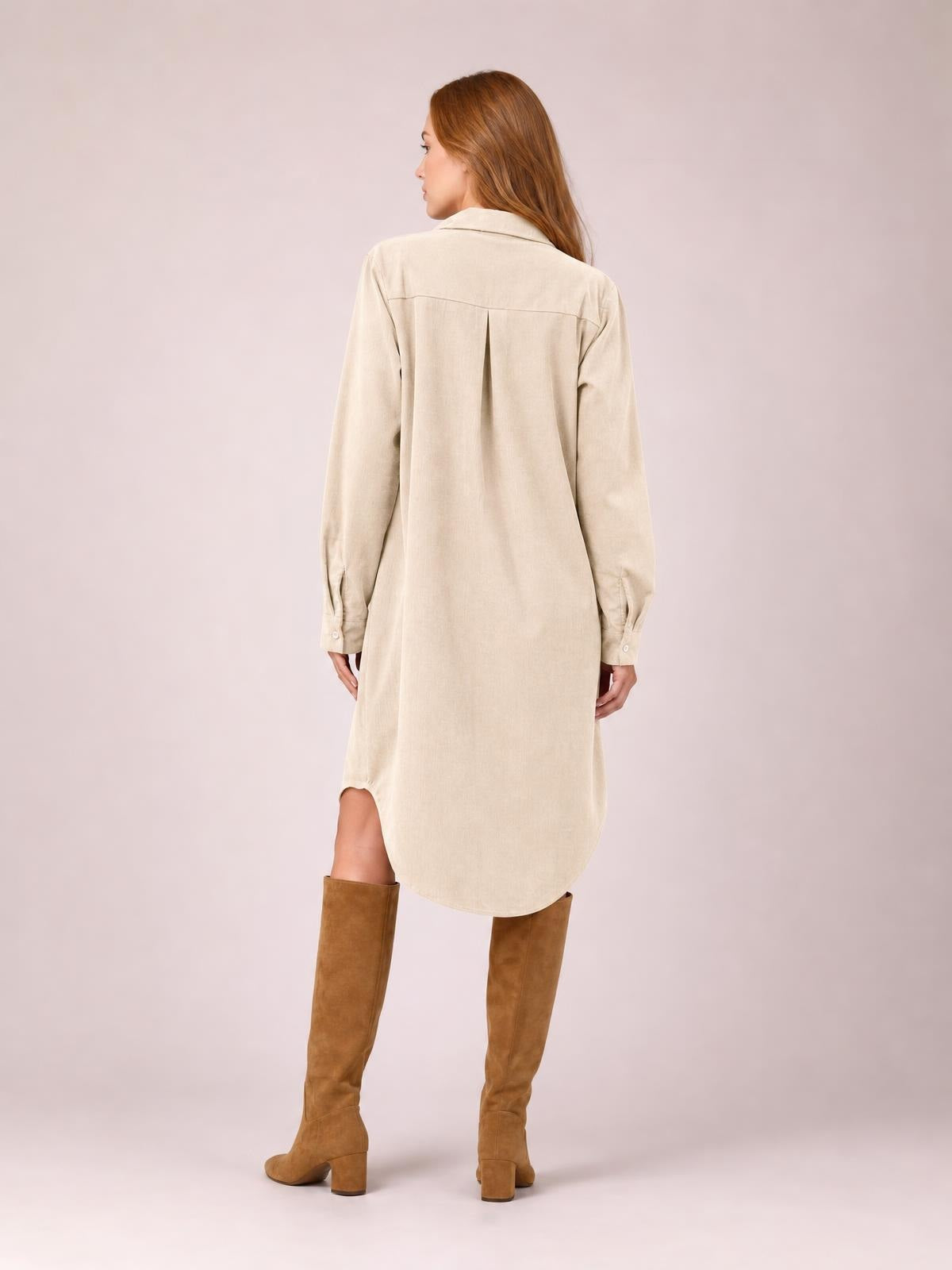 A model stands turned away in a beige corduroy shirt dress with a collared neckline, long sleeves, center-back pleat, and rounded shirt-tail hem.