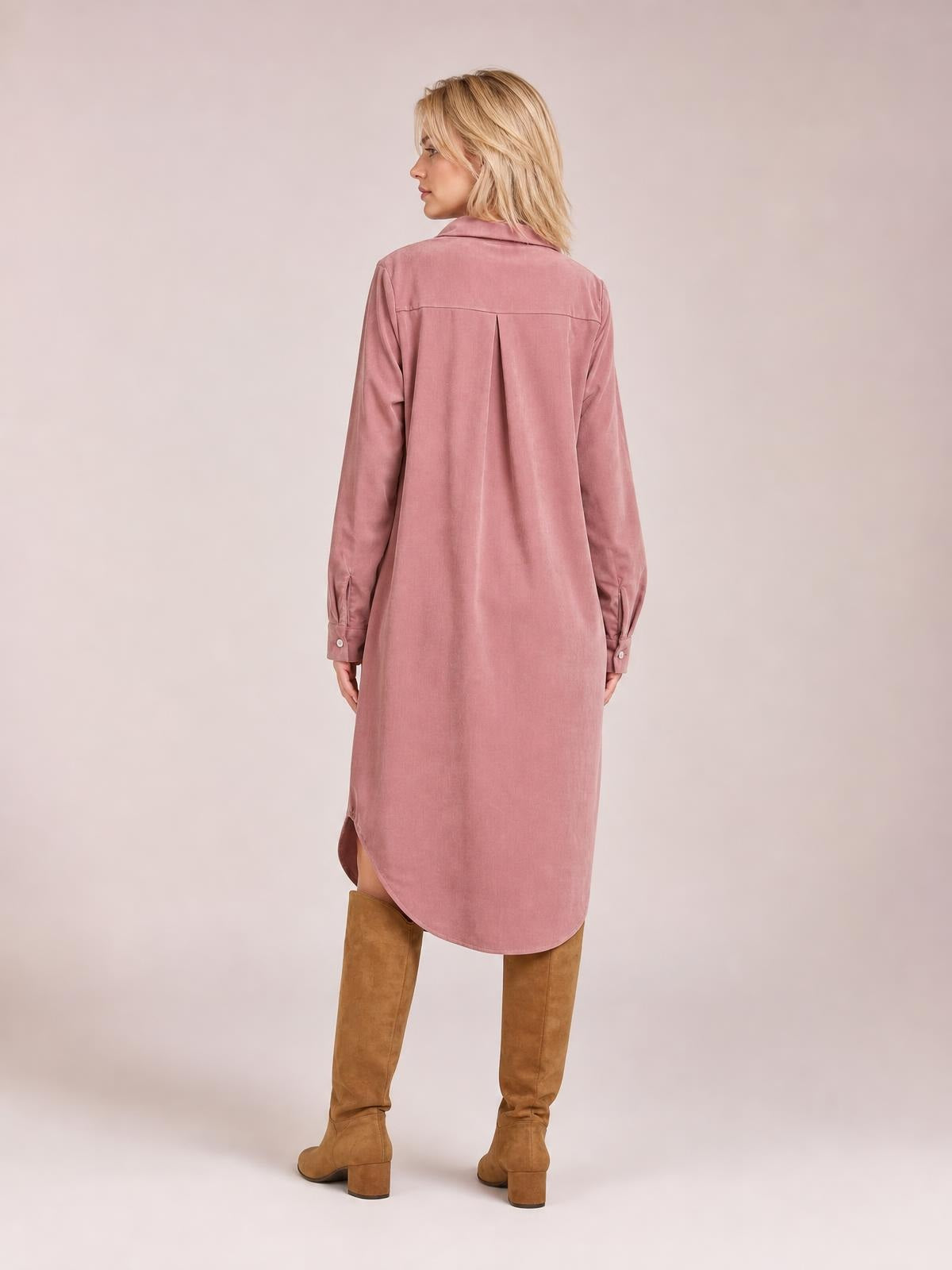 A model stands facing away in a dusty pink corduroy shirt dress with a collared back, long sleeves, center-back pleat, and curved hem.