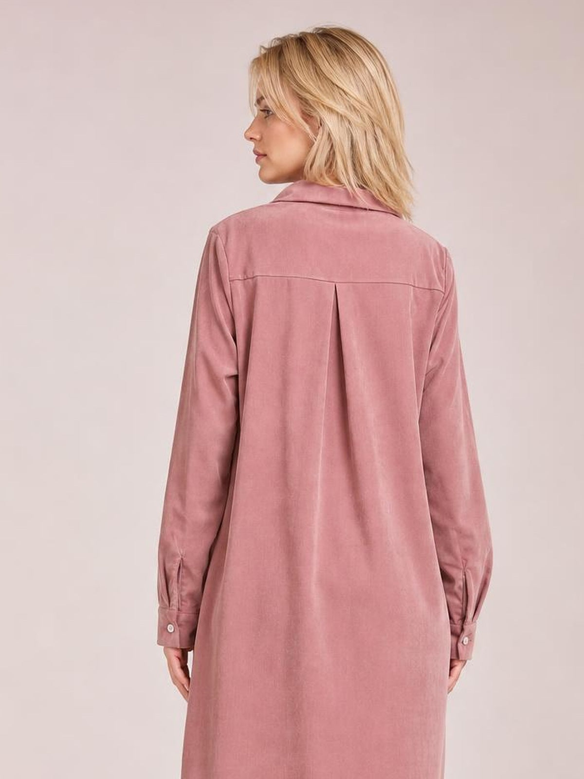 A model faces away in a pink corduroy shirt dress with a collared back, long sleeves, buttoned cuffs, a pleated back yoke, and a relaxed straight silhouette.