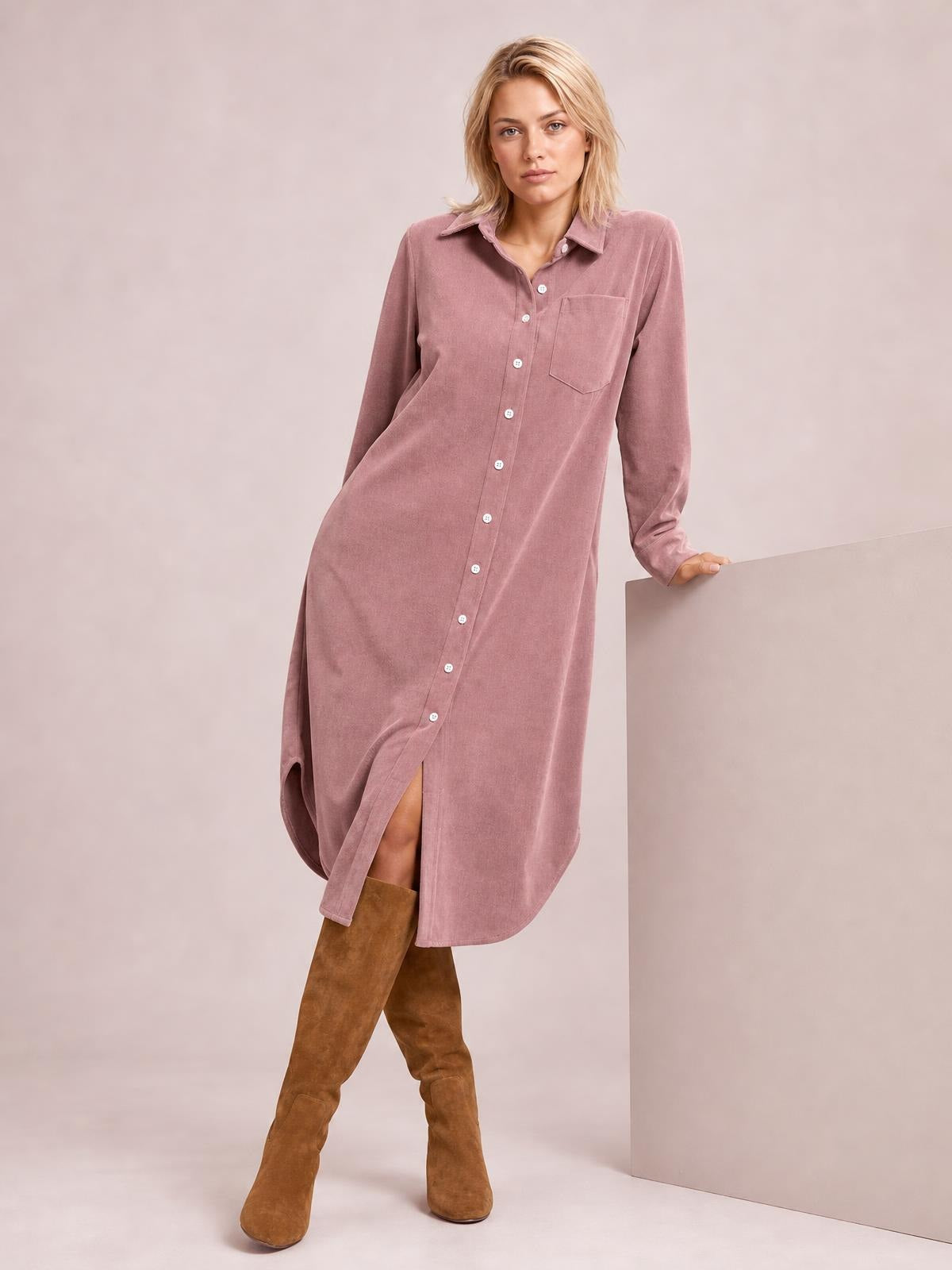 A model leans against a wall in a dusty rose corduroy shirt dress with a button-down front, pointed collar, chest pocket, long sleeves, and relaxed midi silhouette.