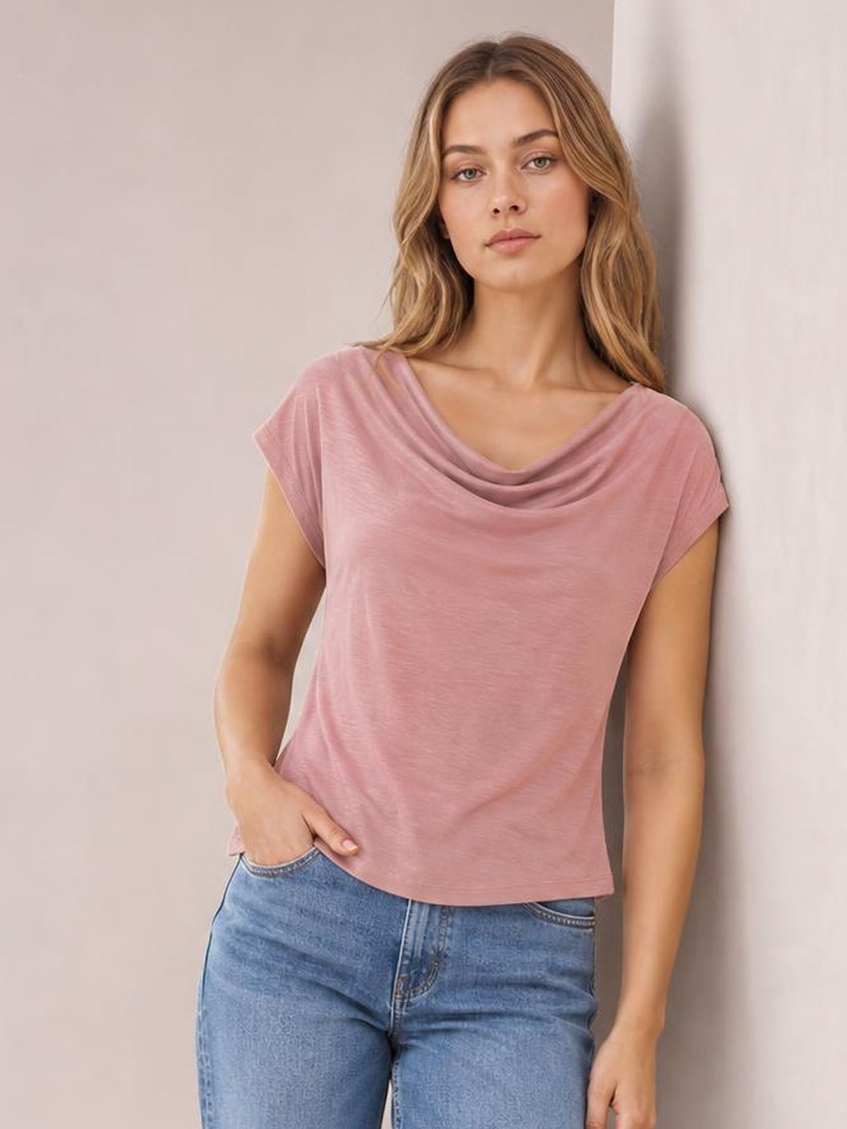 Model wearing a pink cowl neck short sleeve T-shirt with an elegant draped neckline, relaxed silhouette, and straight hem, posing with one hand in her jeans pocket.