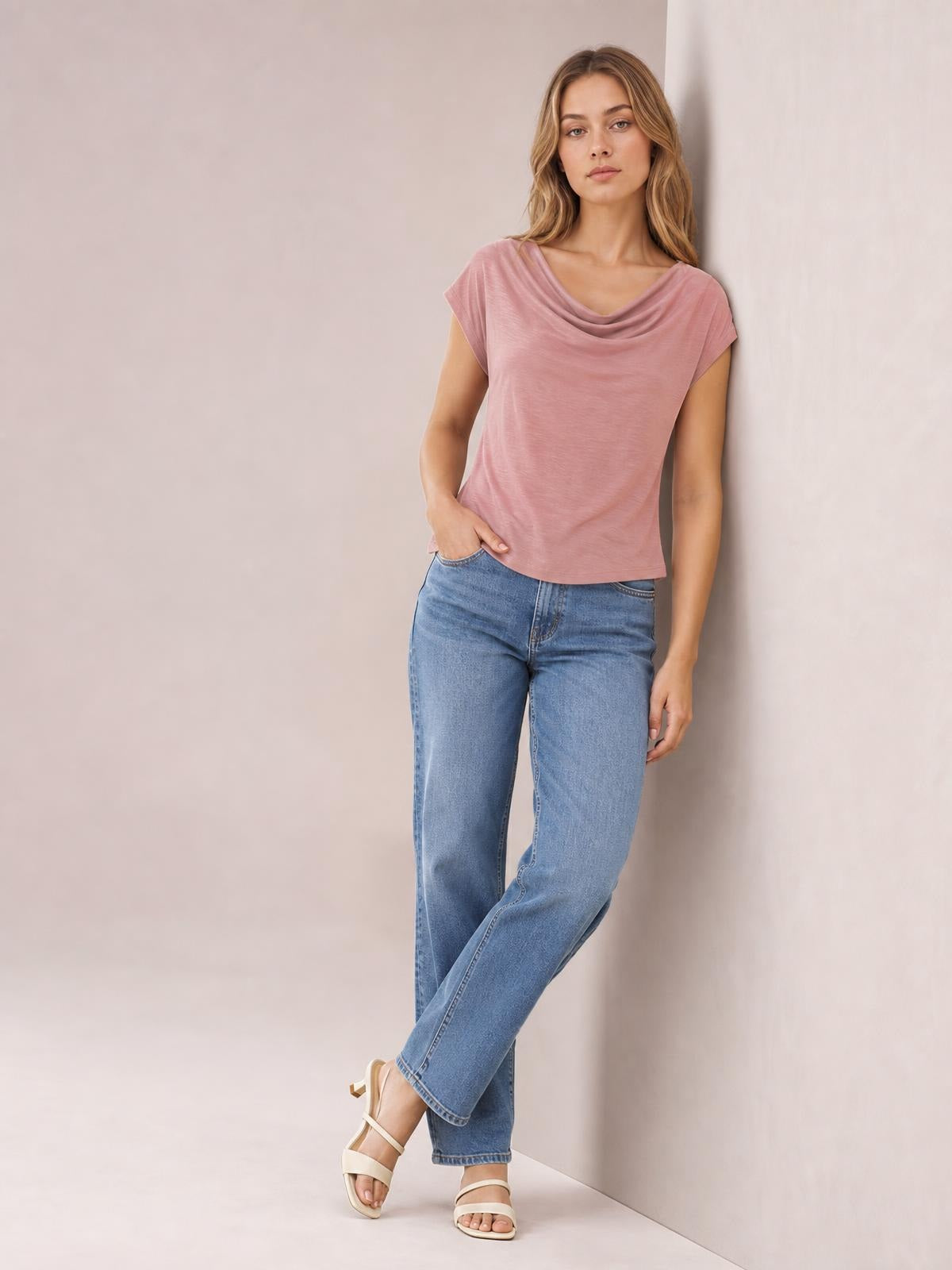 A woman wears a dusty pink cowl neck short sleeve T-shirt with an elegant draped neckline and relaxed silhouette, standing with crossed legs and one hand at her pocket.