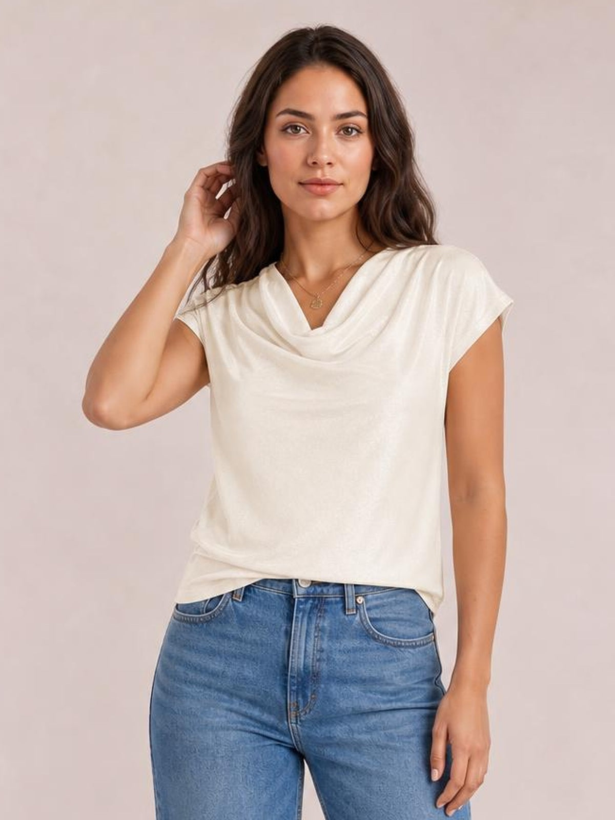 Model poses in a white cowl neck short sleeve T-shirt with a relaxed straight silhouette and softly draped neckline, paired with blue jeans.