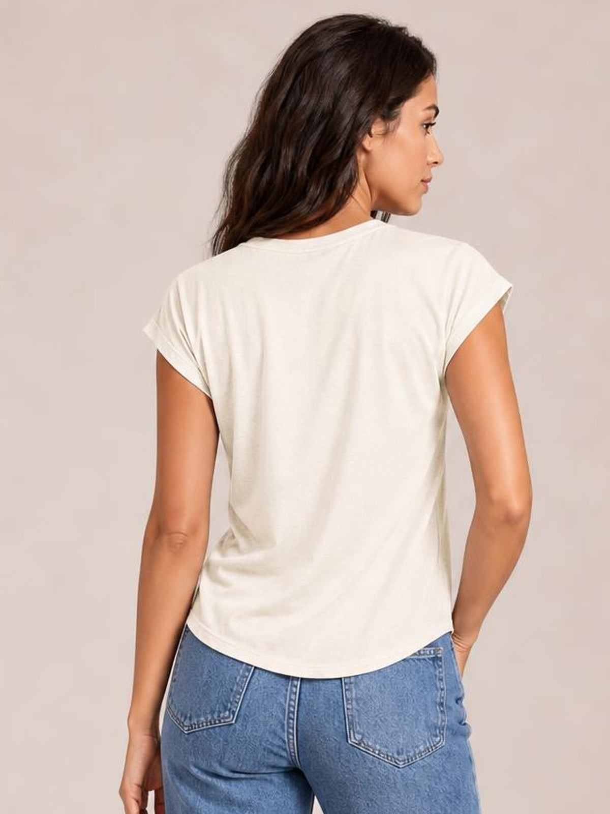 A model wears an opaque white short-sleeve T-shirt with a relaxed silhouette and curved hem, standing turned away with one hand in her pocket.