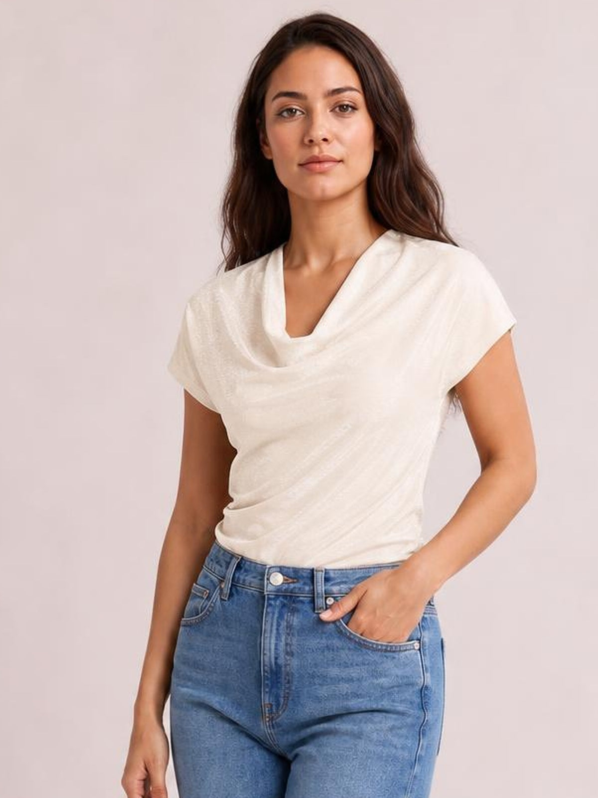 Model wears an ivory cowl neck short sleeve T-shirt with a softly draped neckline, relaxed silhouette, and curved sleeve hems, posing with one hand in her jeans pocket.