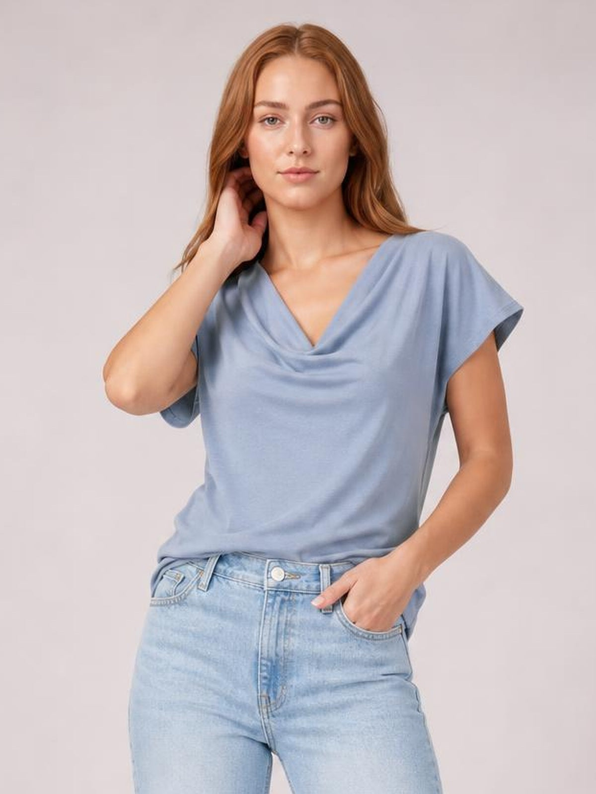 A model poses in a light blue cowl neck short sleeve T-shirt with an elegant draped neckline and relaxed silhouette, paired with jeans.