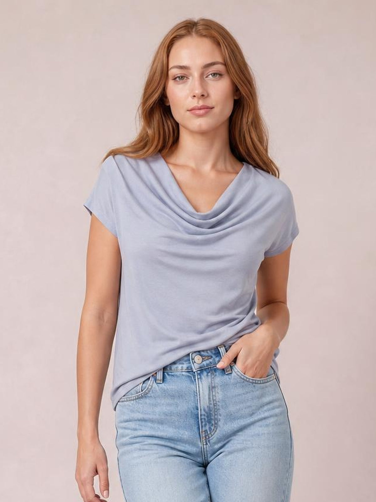 Model wears a light lavender cowl neck short sleeve T-shirt with an elegant draped neckline and relaxed silhouette, standing with one hand in her jeans pocket.
