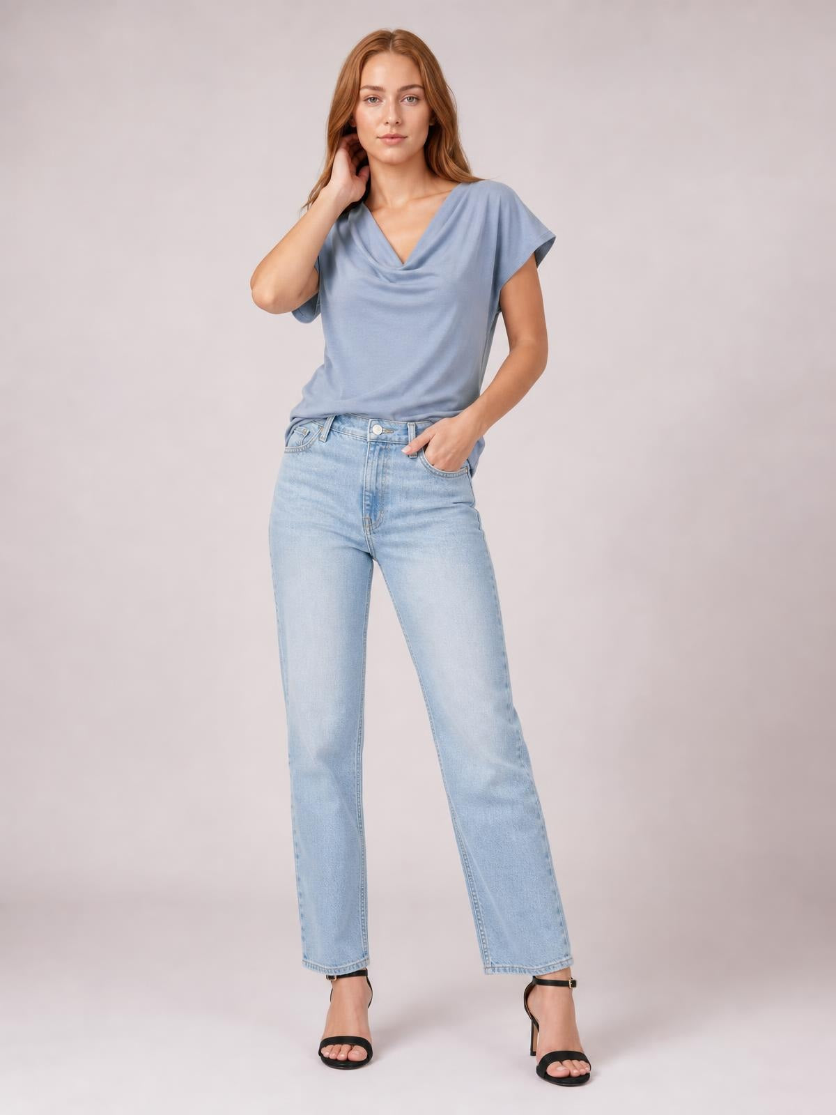 Model poses in a blue cowl neck short sleeve T-shirt with a softly draped neckline and relaxed straight silhouette, paired with light-wash jeans.