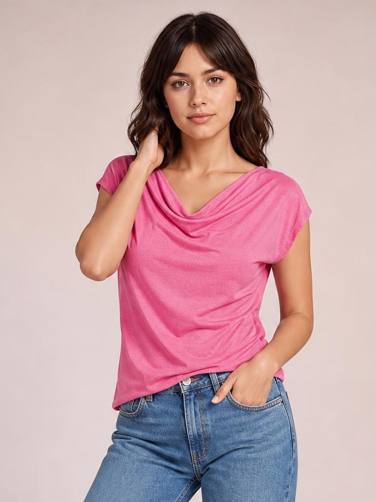 Model wearing a pink cowl neck short sleeve T-shirt with a draped neckline, relaxed silhouette, and gathered side detail, posing with one hand in her jeans pocket.