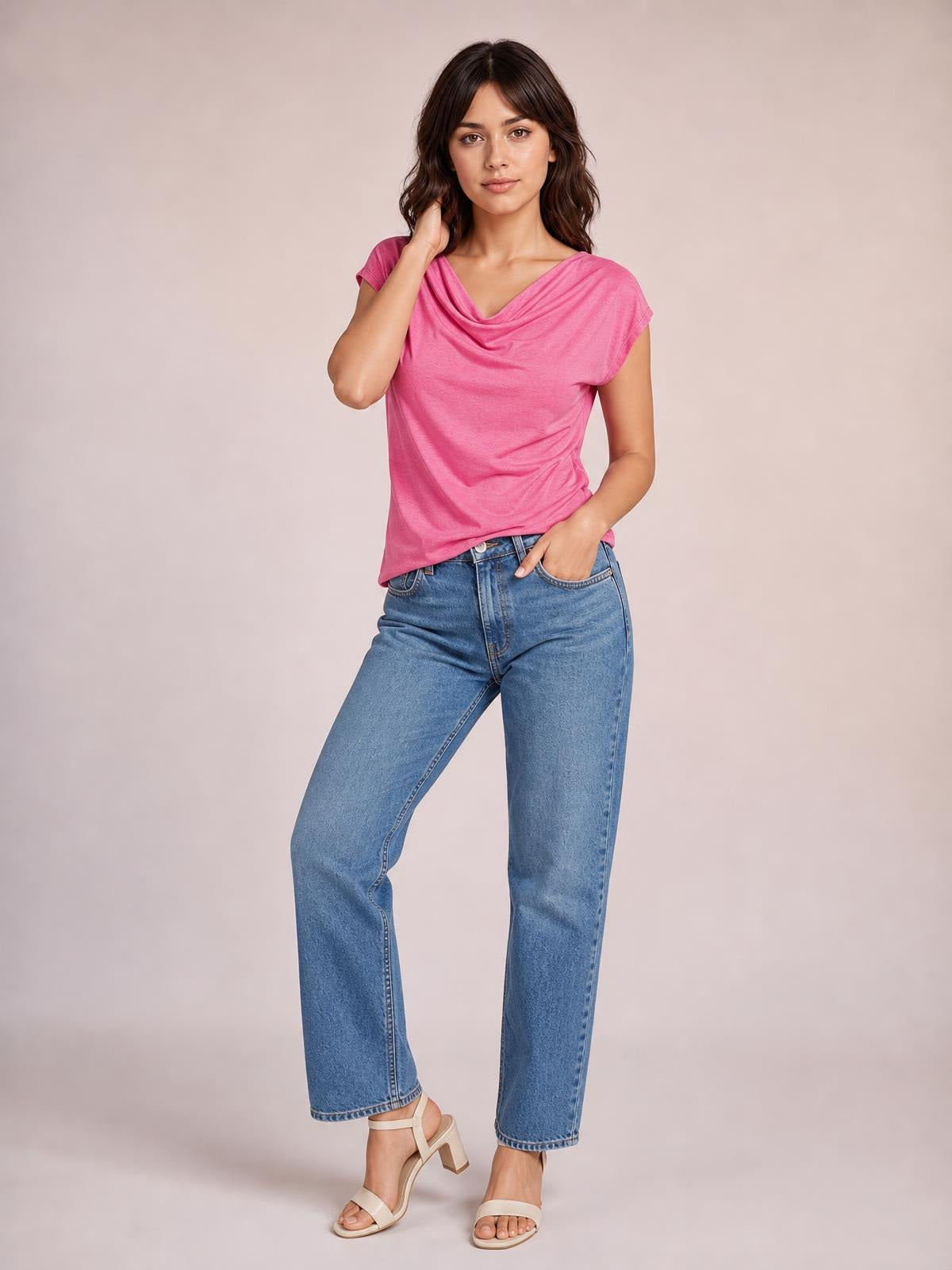 Model wears a bright pink cowl neck short sleeve T-shirt with an elegant draped neckline, relaxed silhouette, and curved hem while posing with one hand in her jeans pocket.