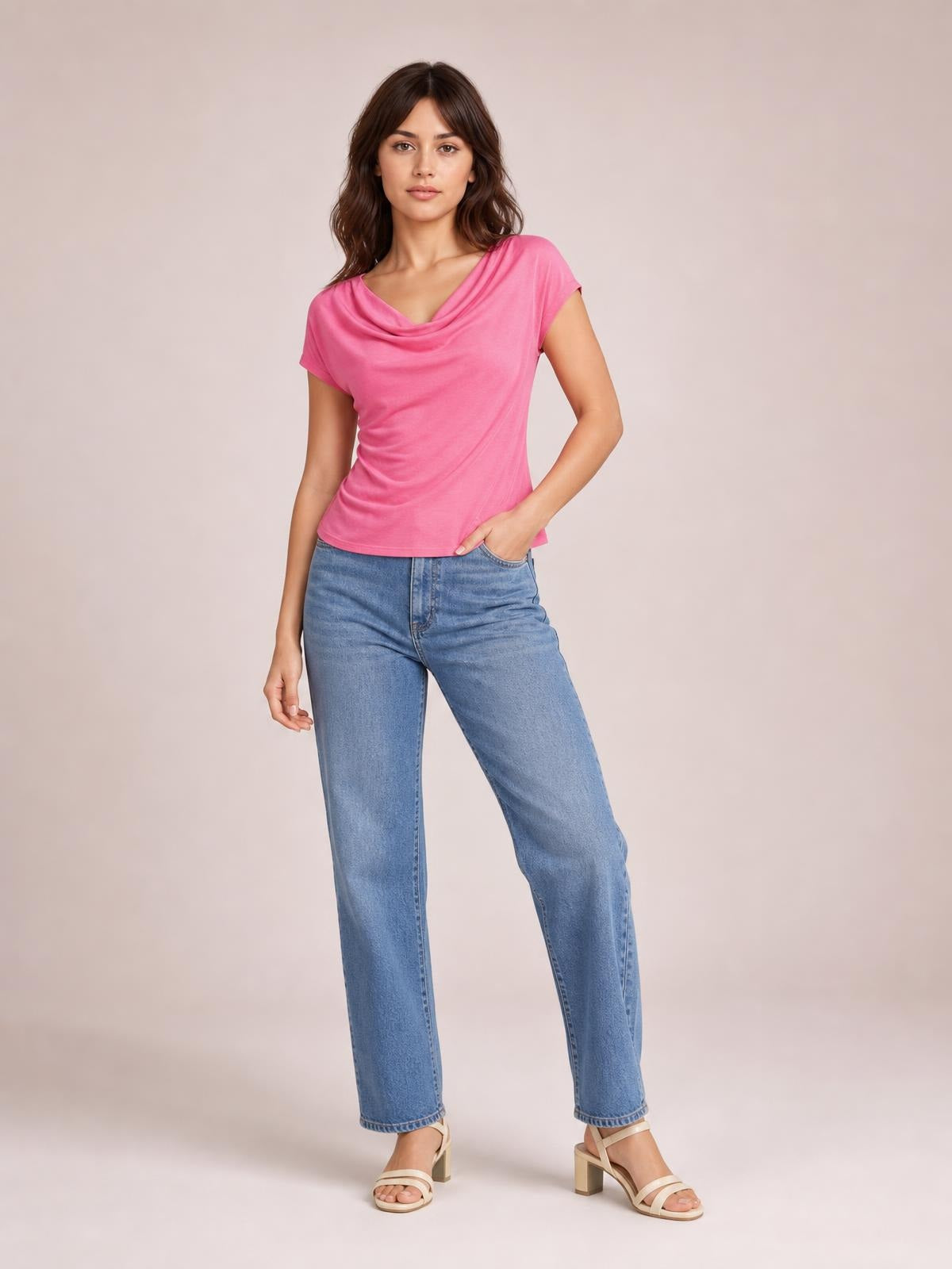 Model poses in a bright pink cowl neck short sleeve T-shirt with a draped neckline and fitted silhouette, paired with light-wash jeans.