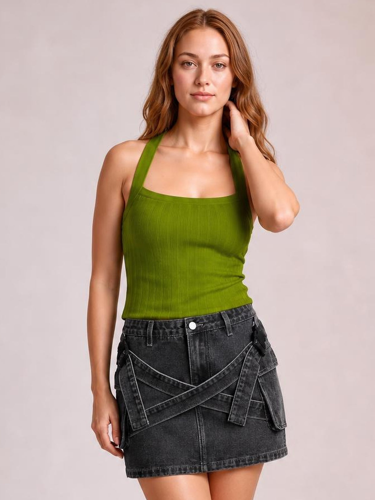 A model poses in a dark charcoal denim mini skirt with a structured straight silhouette, front pockets, and bold criss-cross waist detailing.