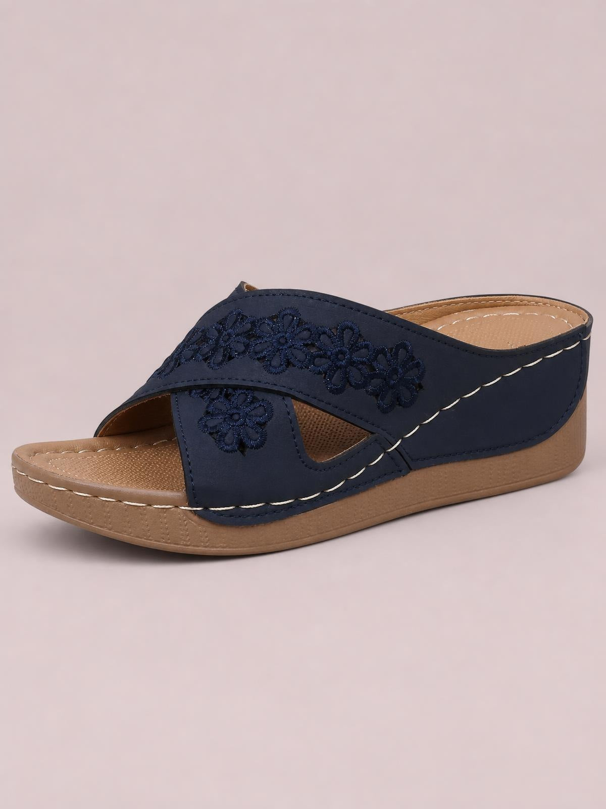 Navy crisscross flower wedge sandal with open toe, decorative floral straps, tan cushioned footbed, low wedge heel, and contrast stitching.
