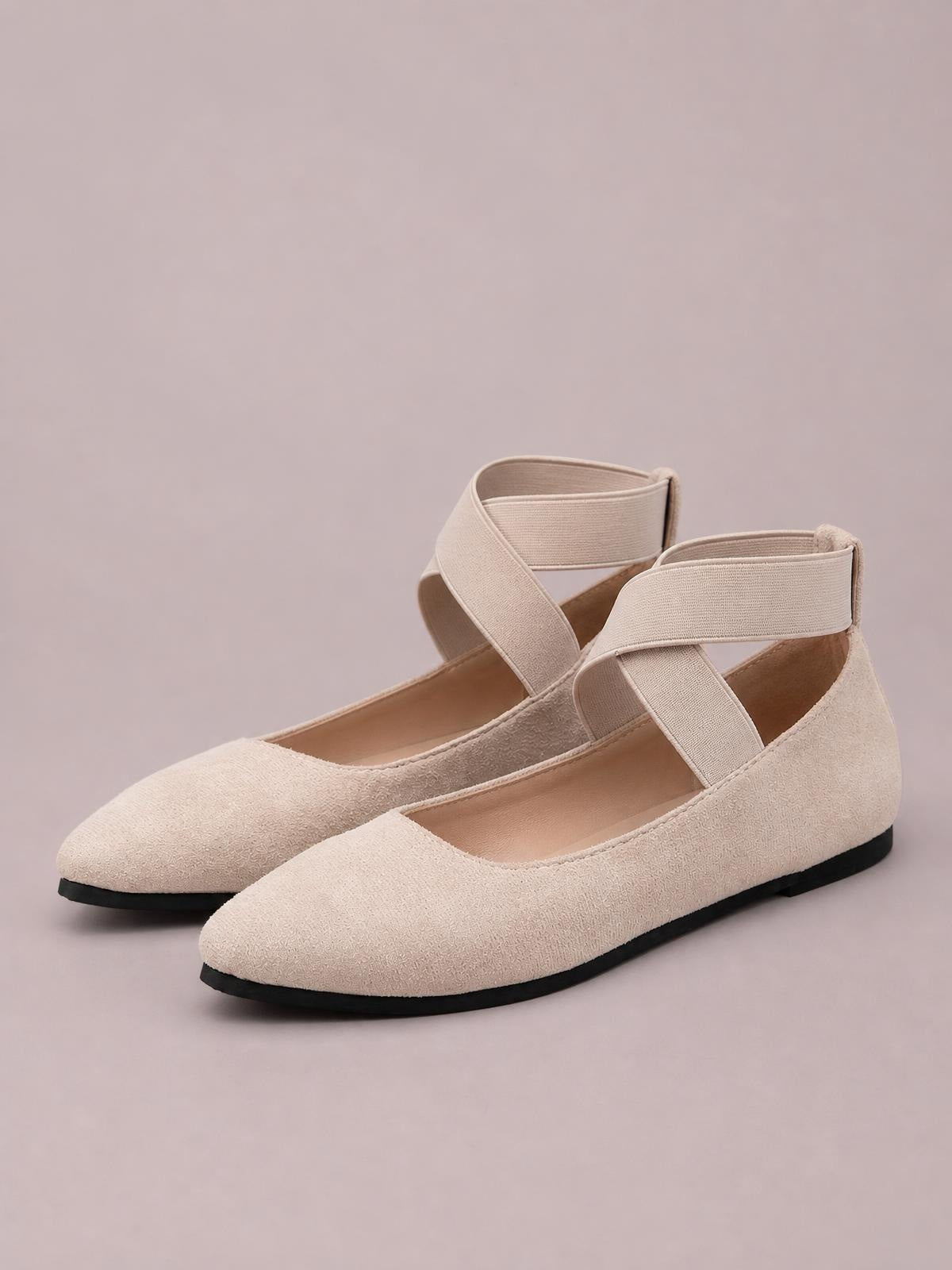 A pair of beige suede flats features crisscross ankle straps, tapered toes, and low-profile black soles in a close-up product view.