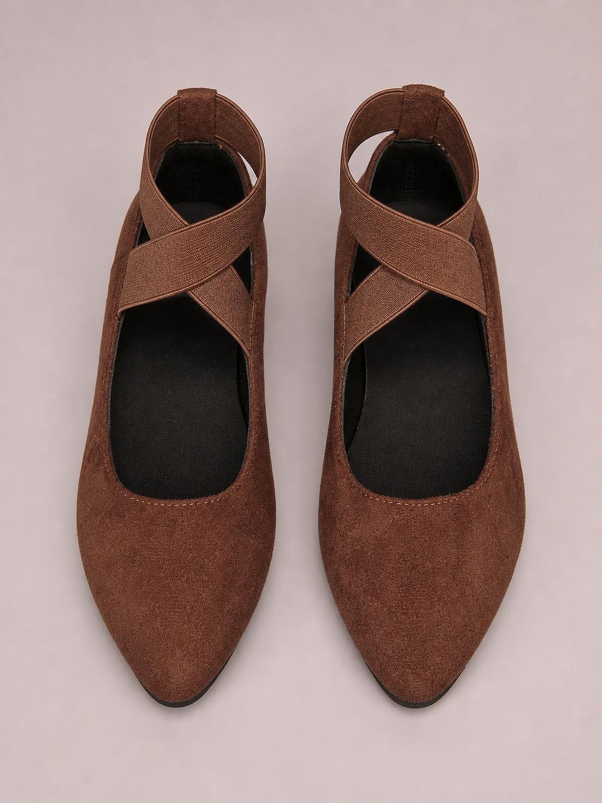 Brown suede point-toe flat slip-ons featuring wide crisscross straps, open sides, black insoles, and slim rubber soles.