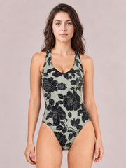 Model stands with arms at her sides in a fitted sage-green one-piece swimsuit with bold black floral print, scoop neck, and wide shoulder straps.