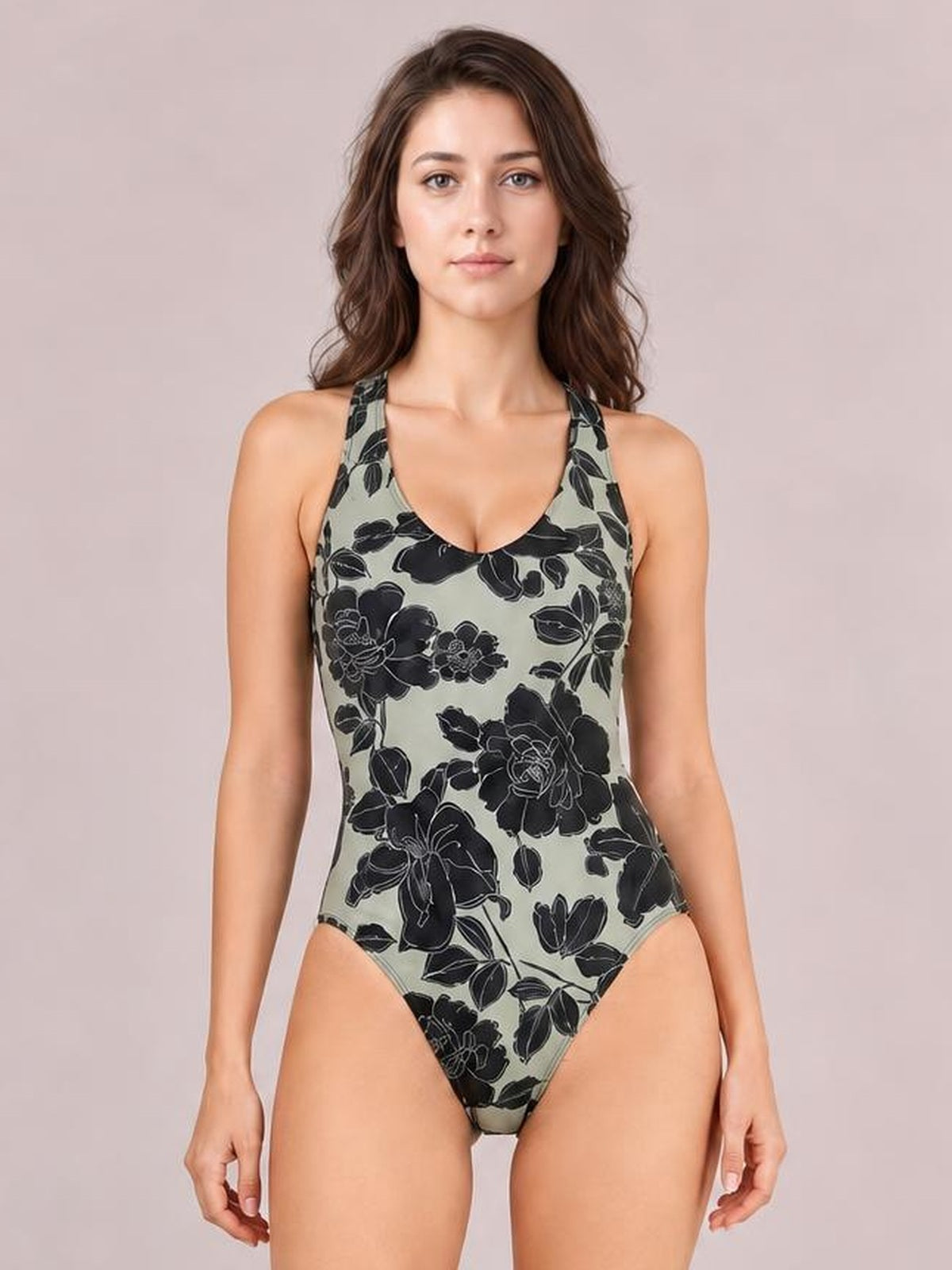 Model stands with arms at her sides in a fitted sage-green one-piece swimsuit with bold black floral print, scoop neck, and wide shoulder straps.