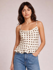 Model wearing an ivory polka dot cami with thin straps, a fitted ruched silhouette, and a playful scalloped ruffled hem, paired with jeans.