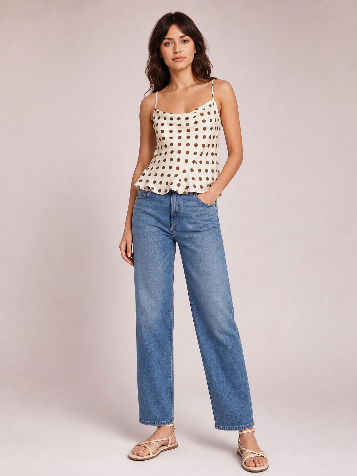 Model wears an opaque cream polka dot cami with thin straps, a softly draped neckline, fitted silhouette, and playful ruffled hem, paired with relaxed jeans.