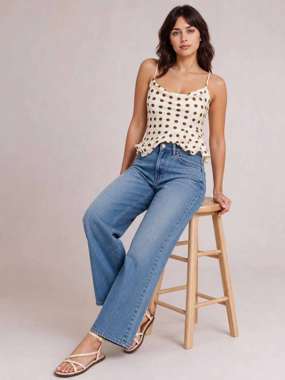 A seated model wears an ivory polka dot cami with thin straps, a gently ruched fitted bodice, and a playful ruffled hem over wide-leg jeans.