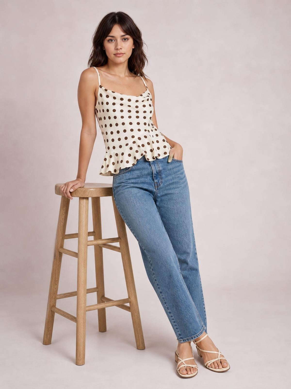 A model poses in an ivory polka dot cami with thin straps, a fitted ruched bodice, and a flared ruffled hem while leaning against a stool.