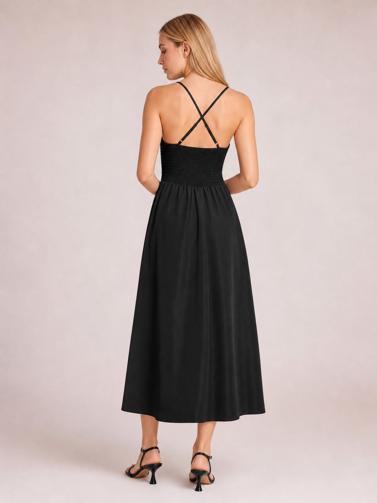 A model stands in a black midi dress with spaghetti straps, a crisscross back, smocked bodice, defined waist, and flowing A-line skirt.