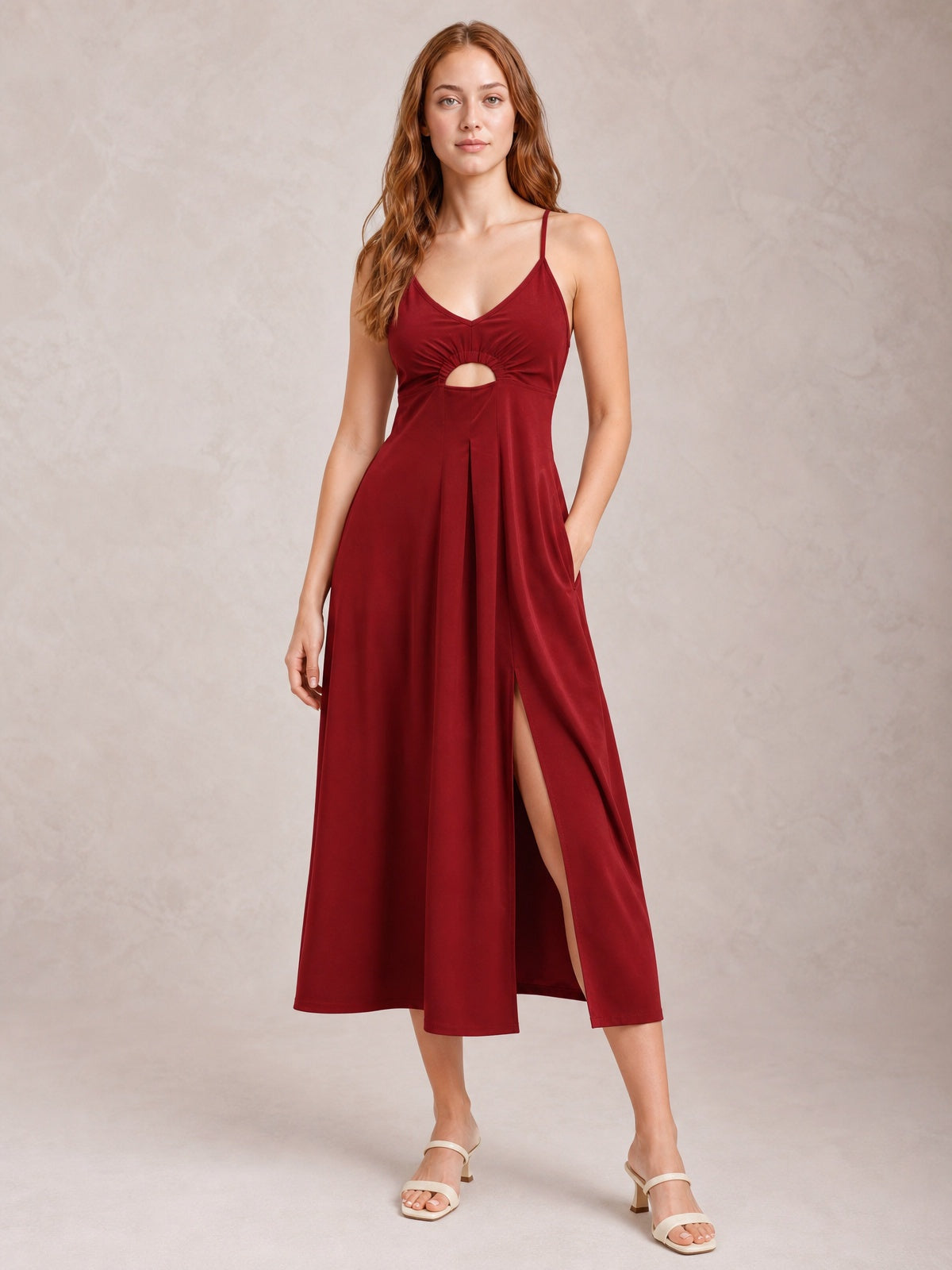 Model stands in a burgundy spaghetti-strap midi dress with a gathered bodice, front cutout, defined waist, flowing A-line skirt, and side slit.