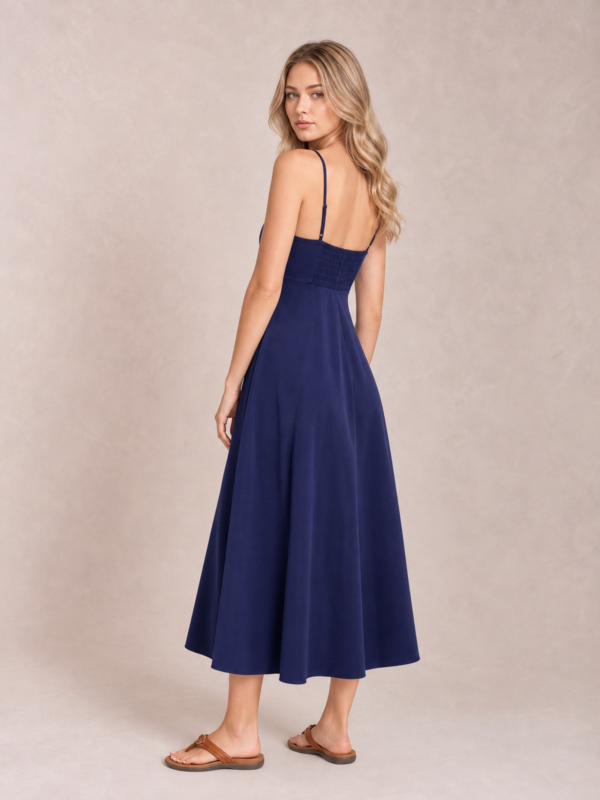 Model poses in a navy crisscross smocked spaghetti strap dress with an open back, defined waist, and flowing midi-length skirt.
