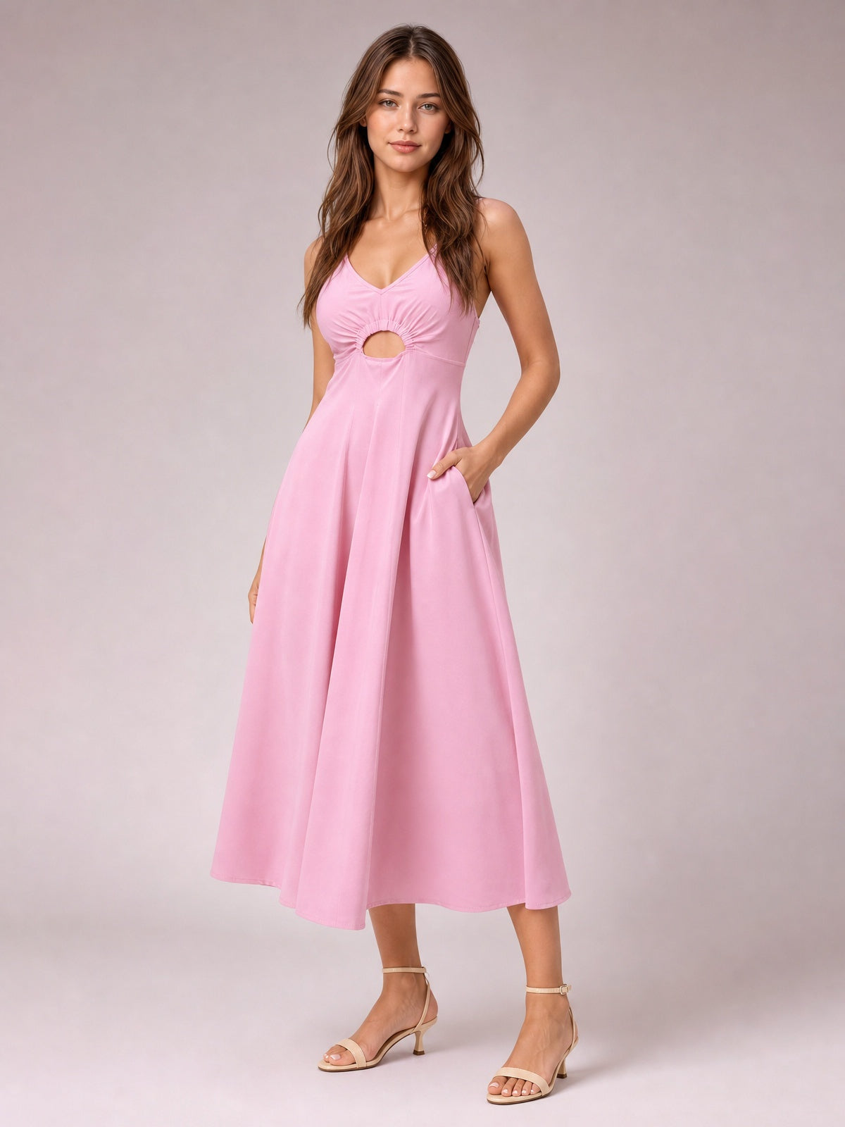 A model poses in a pink spaghetti-strap dress with a gathered bodice, front cutout, defined waist, side pockets, and a flowing midi-length skirt.