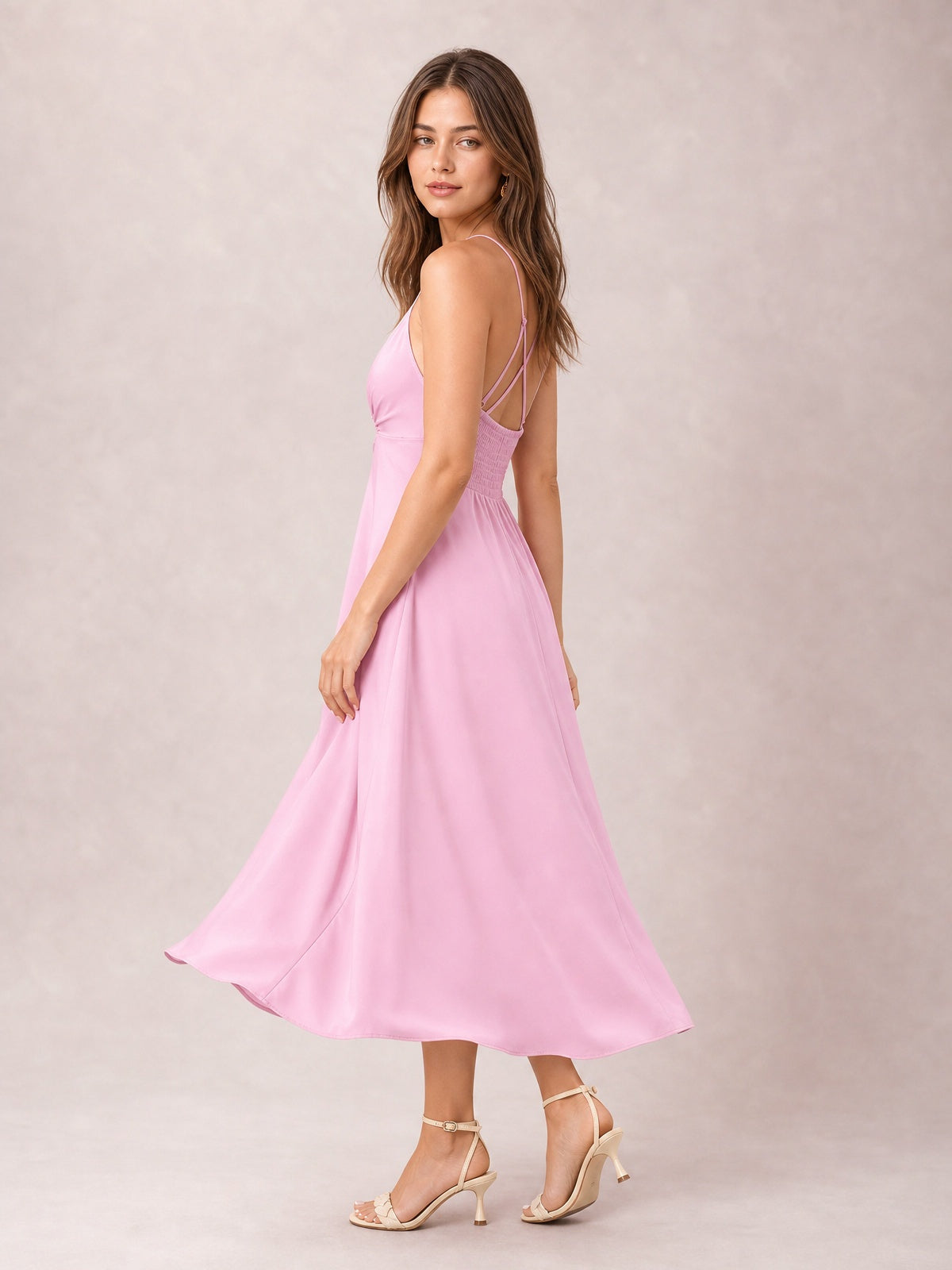 A model turns in a pink crisscross smocked spaghetti strap dress with an open back, defined waist, and flowing midi-length skirt.