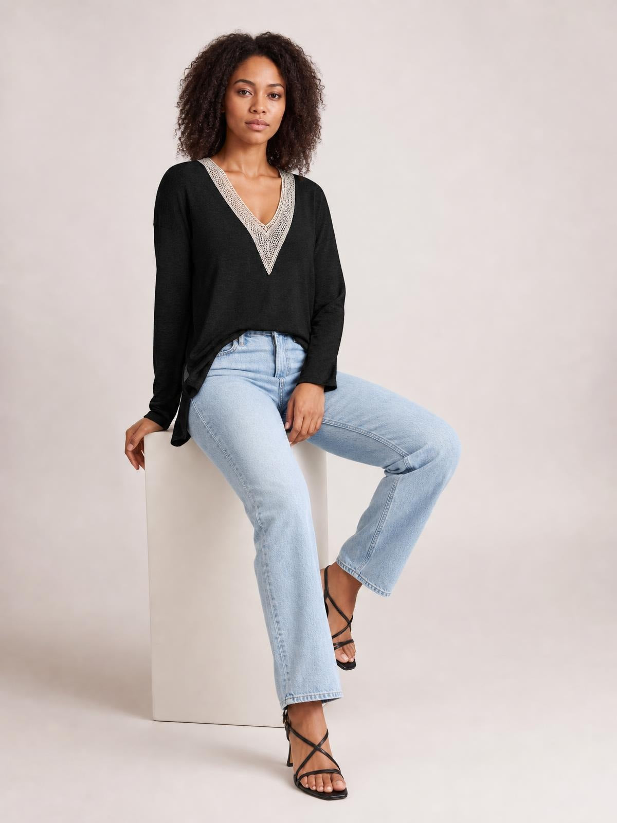 Model seated in a black long-sleeve T-shirt with a contrasting crochet V-neck, relaxed silhouette, and side slit hem.