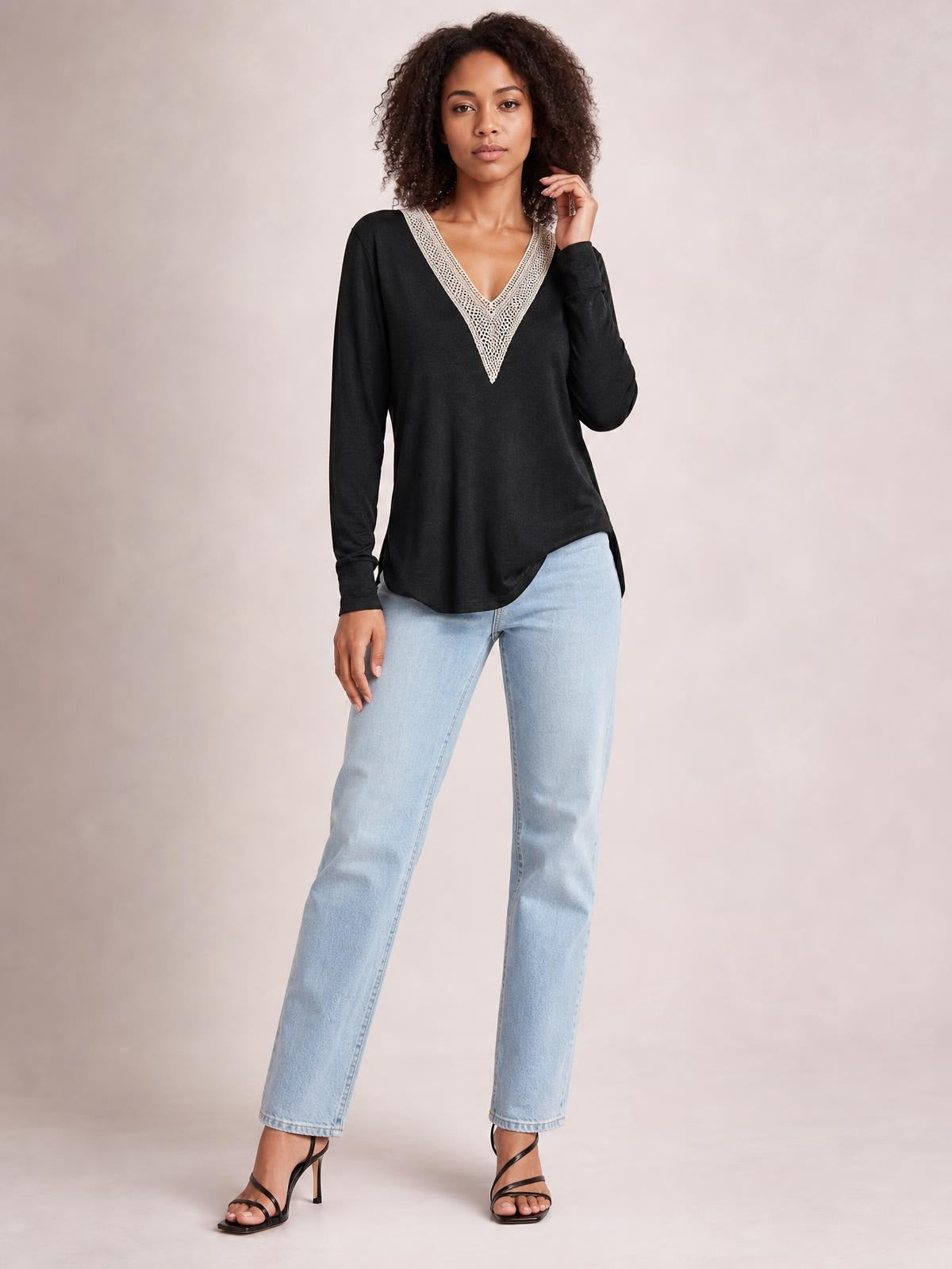 Model wears a black long-sleeve T-shirt with a contrasting crochet V-neck, relaxed silhouette, curved hem, and side slit while posing with one hand near her hair.