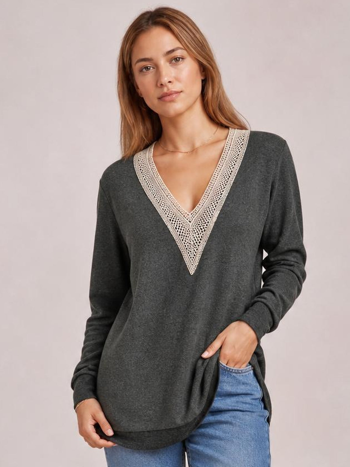 A model wears a charcoal long-sleeve T-shirt with a contrasting crochet V-neck, relaxed silhouette, curved hem, and side slit while standing with one hand in her jeans pocket.