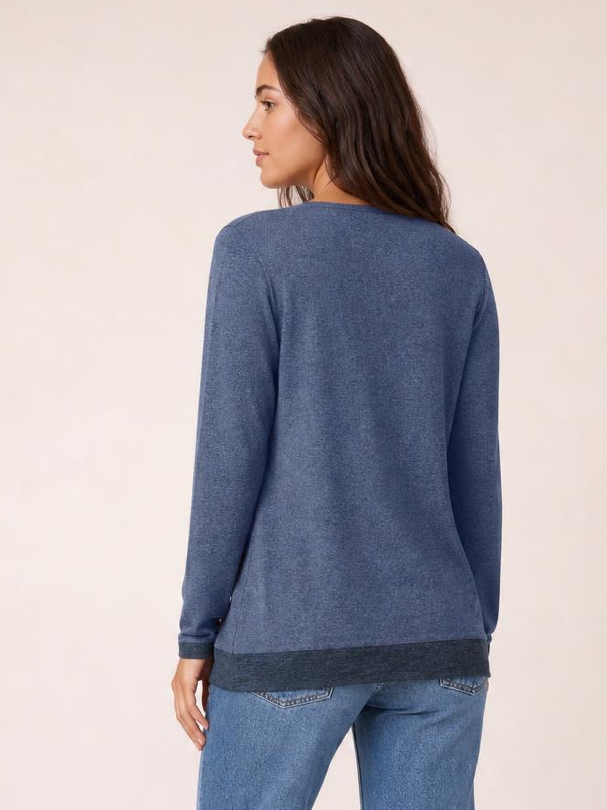 Woman shown from the back in a blue long-sleeve T-shirt with a relaxed silhouette and contrasting dark hem, standing with one hand at her side.