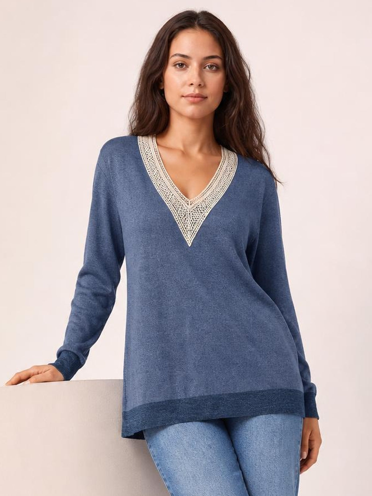 A model wears a relaxed blue long-sleeve T-shirt with a cream crochet contrast V-neck, dark hem band, and gently flared silhouette, resting one hand on a ledge.