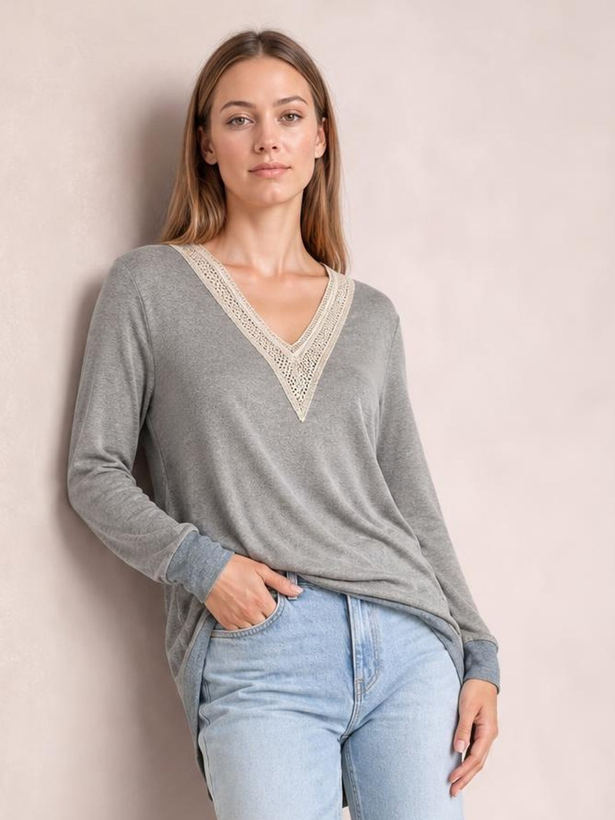 Model wears a gray long-sleeve T-shirt with a crochet contrast V-neck, relaxed silhouette, and slit hem while leaning with one hand in her jeans pocket.