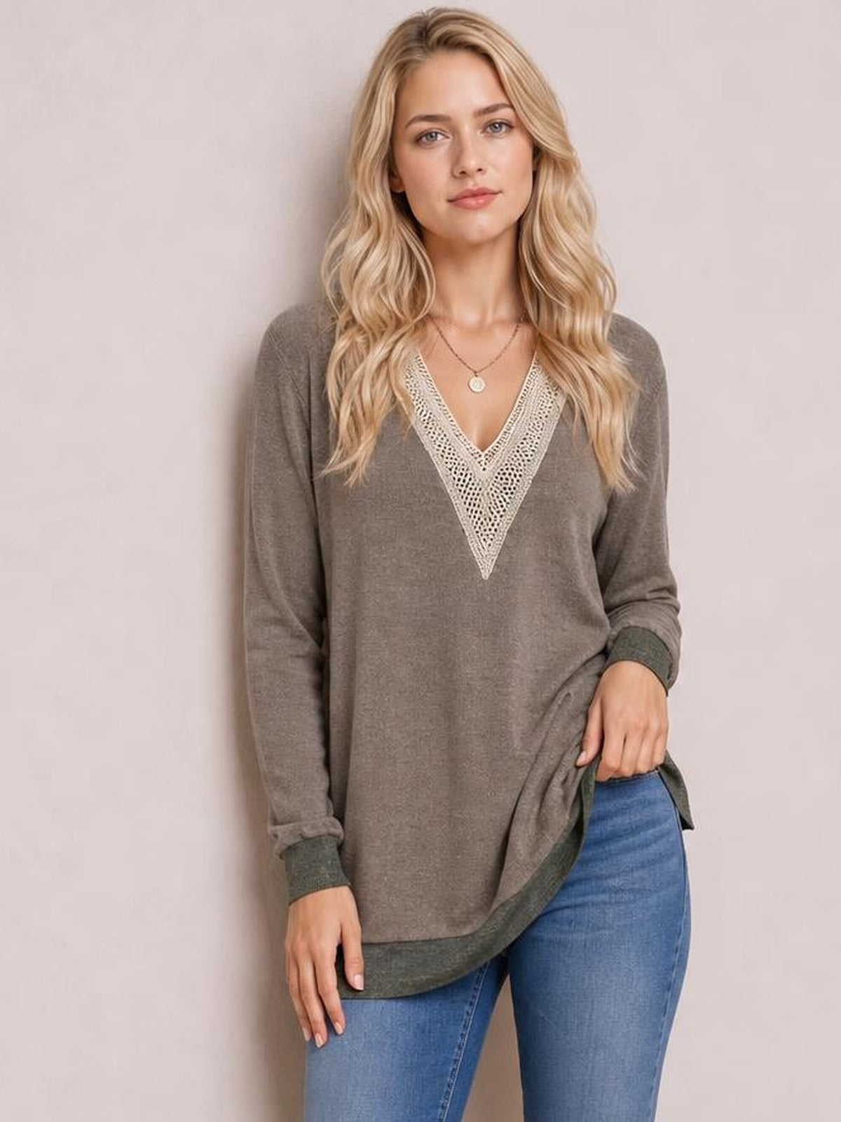 Model wears a taupe long-sleeve T-shirt with a contrasting crochet V-neck, relaxed silhouette, dark cuffs, and side slit while lifting the hem.