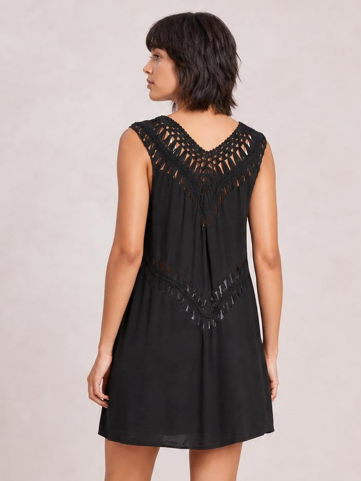 A model stands facing away in a black beach cover-up dress with a loose A-line silhouette, crochet-detail cutout yoke, and decorative openwork waist panel.