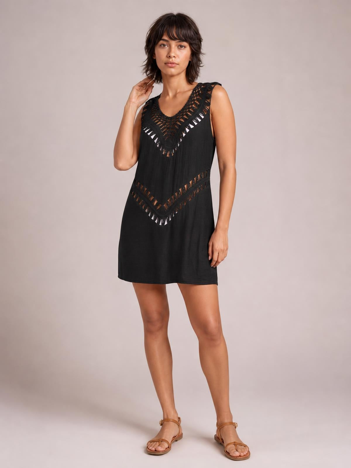 A model poses in a black beach cover up dress with crochet-detail cutout panels at the neckline and waist, a sleeveless design, and relaxed short silhouette.