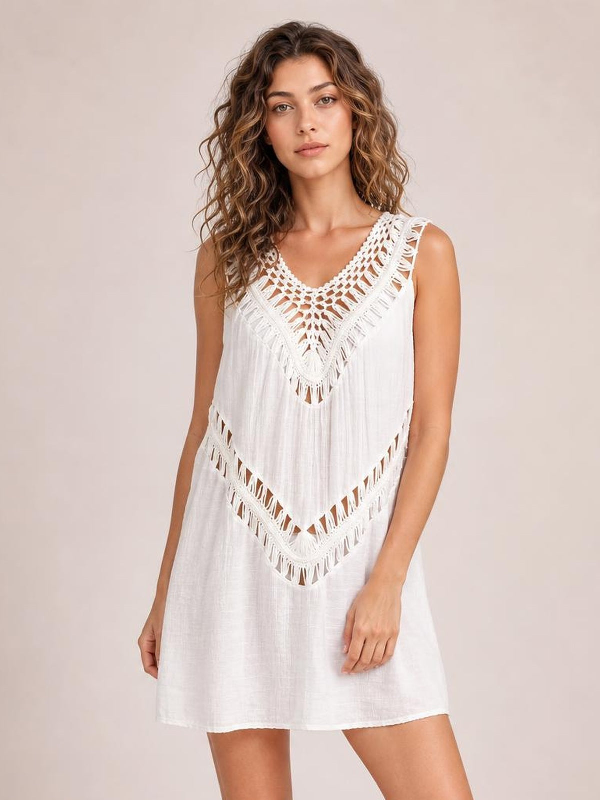 Model stands with arms relaxed in a white crochet detail beach cover up dress featuring a loose silhouette, sleeveless cutout neckline, and fringe trim.