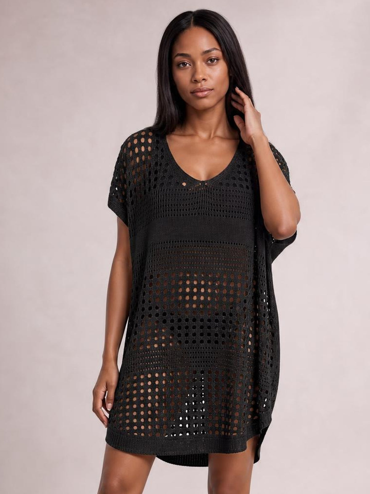 Model poses with one hand in her hair wearing a black crochet knit beach cover up dress with a loose silhouette, V-neck, and openwork cutout construction.