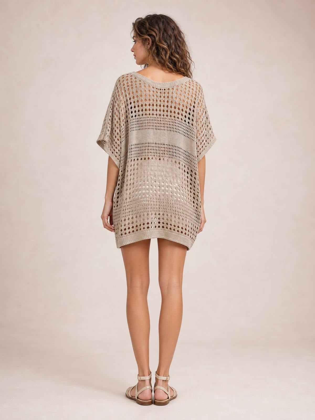 Model shown from the back in a beige crochet knit beach cover up dress with openwork cutout pattern, loose oversized silhouette, short sleeves, and curved hem.