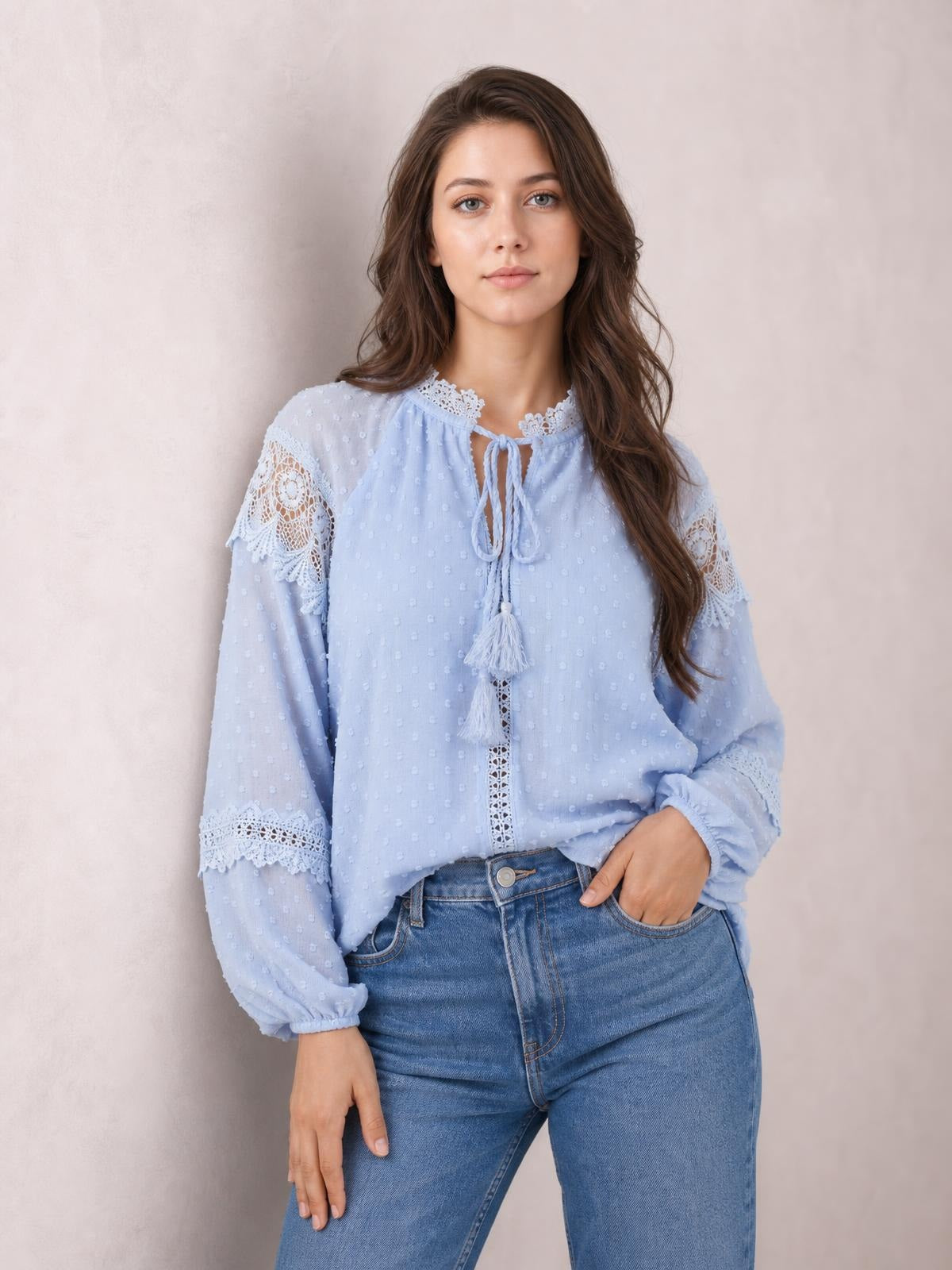 A model poses in a light blue crochet blouse with a tassel tie neckline, long puff sleeves, lace trim, and a relaxed, semi-sheer silhouette.