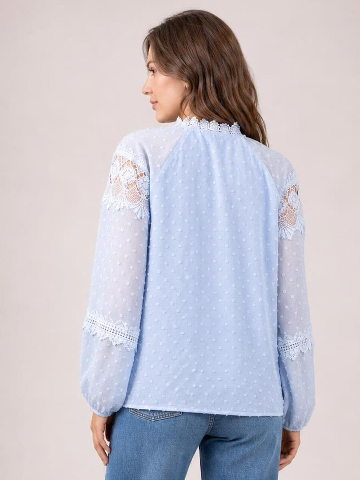 A model turned away wears a light blue blouse with a relaxed silhouette, textured dotted fabric, long sheer sleeves, and crochet lace trim at the collar and cuffs.
