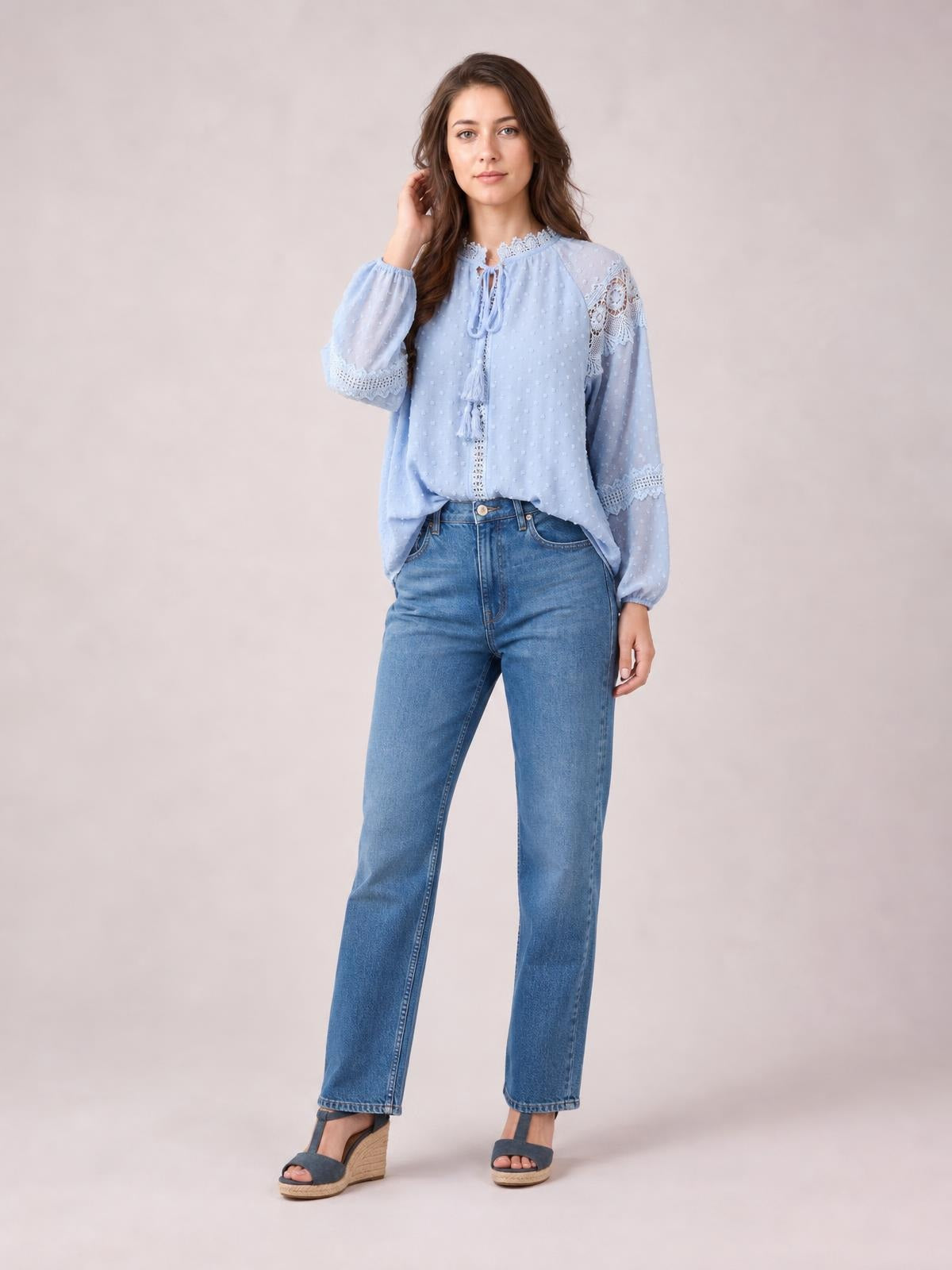 A model poses in a light blue crochet tassel tie-neck blouse with long sheer sleeves, lace-trimmed cuffs, and a relaxed, flowing silhouette.